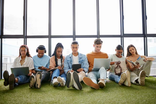 seven young people of different races and genders sitting beside each other on a green carpet in front of a window on their laptops, tablets, and phones