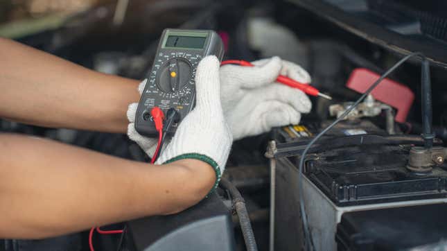 Close-up of mechanic holding voltmeter to check voltage car battery energy problem for service maintenance. Car mechanic noting repair parts during open car hood engine repair unrecognisable