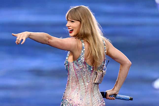 Taylor Swift with her back turned to the camera but her face turned to her left, smiling, and pointing with her left hand, her right hand is by her side holding her mic