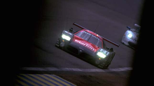 A photo of a 1997 Nissan R390 GT1 racing at Le Mans, viewed from the front at an angle with its lights on.