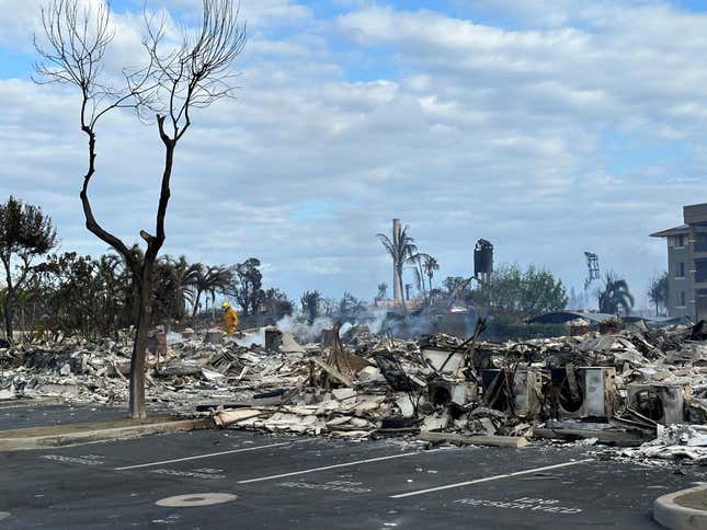 A firefighter puts out hot spots in a parking lot covered in burnt debris. A charred tree stands in the foreground.