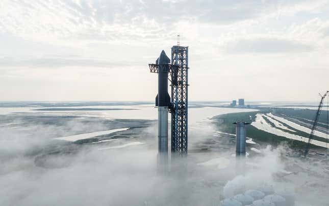 A photo of the Starship rocket docked at its launchpad, towering over the landscape and surrounded by clouds.