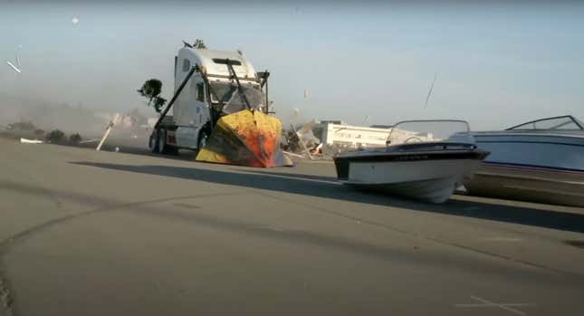 A semi truck with a pointed nose welded to its front and no trailer speeds towards two boats sitting on a tarmac. 