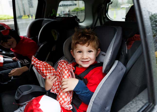 A child in a red parka sits in a car seat holding something wrapped in red and white checked paper