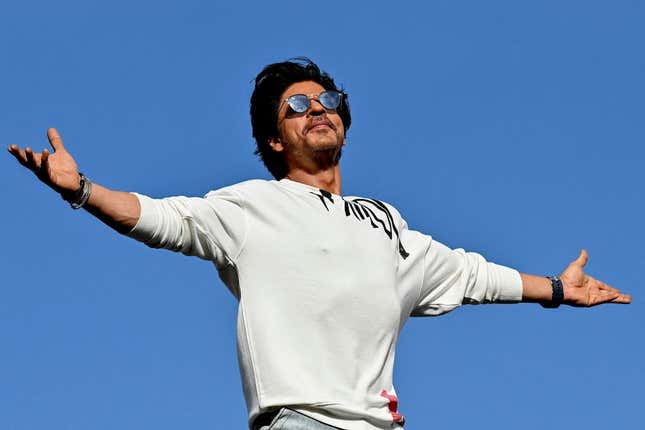 Shahrukh Khan, wearing sunglasses and a sweatshirt, spreads his arms wide in his signature move.