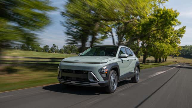 A greenish Kona driving in front of trees