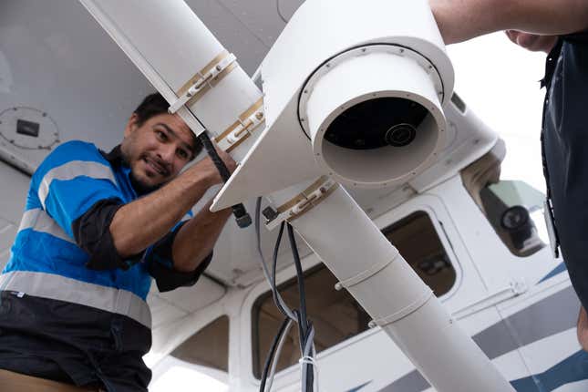 a man wearing a blue and grey jacket fixing wiring on the wing of a plane