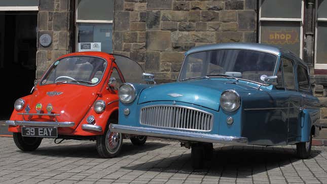 A photo of two vintage three-wheeled cars parked on a cobble street. 