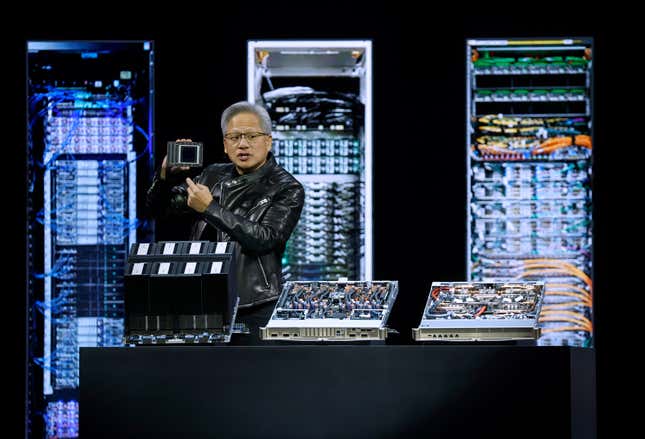 Jensen Huang holding up a chip behind a table with other chip platforms in front of a backdrop with images of server racks