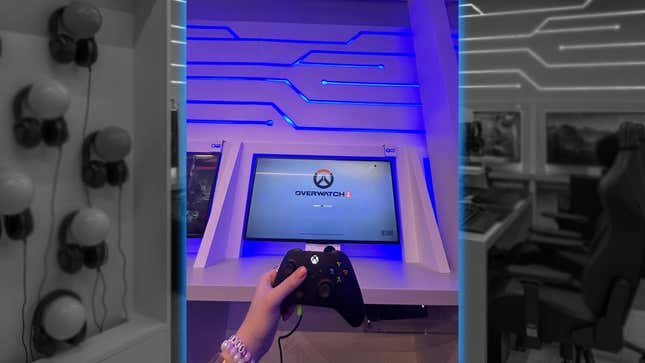 A hand holds an Xbox controller in front of a display showing the Overwatch 2 start screen. 