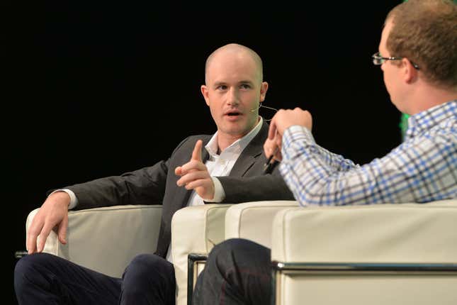 Coinbase CEO Brian Armstrong sitting in a chair, raising a finger, while talking to a moderator at a conference