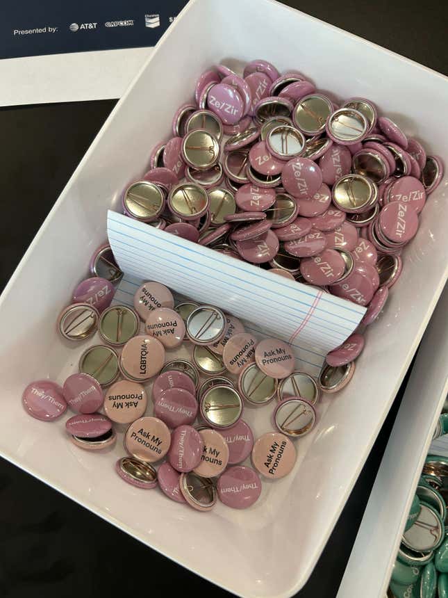 A container offers an assortment of pronoun pins, with options including "They/Them," "Ze/Zir," and "Ask My Pronouns."