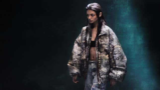 A model wears a puffer jacket featuring the Elden Ring map printed on it, unzipped to reveal her midriff and jeans. 