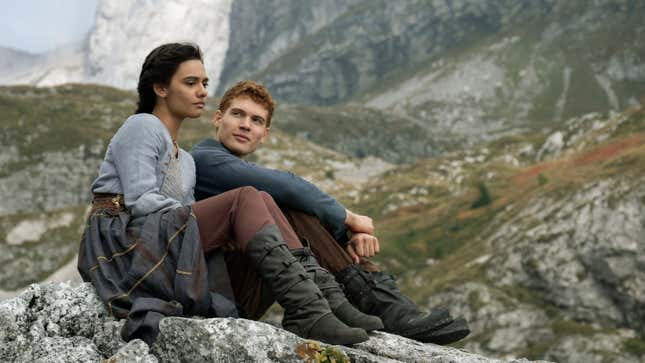 Two young people, a man and a woman, sit together on a rocky hillside in a scene from Wheel of Time.