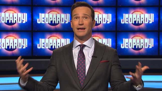 A boring man in a boring suit standing in front of a wall of Jeopardy! monitors.