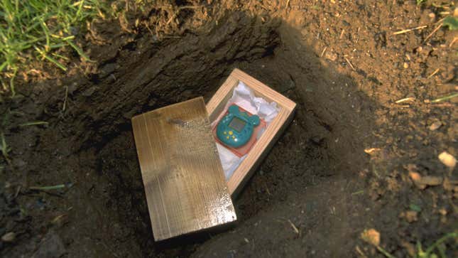 A Tamagotchi appears in a wooden coffin inside a hole in the dirt.