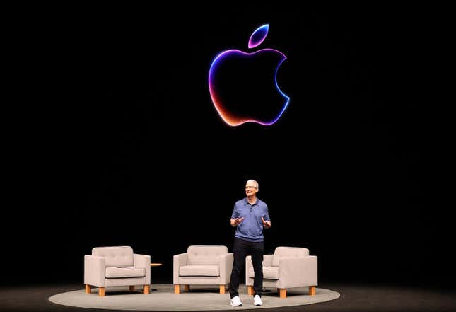 Tim Cook standing on stage in front of three grey chairs, a black backdrop is an Apple logo outlined in neon colors is behind him