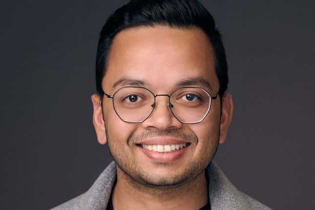 Primer plano de Viswa Colluru con gafas y sonriendo con un cuello de chaqueta gris claro delante de un fondo gris