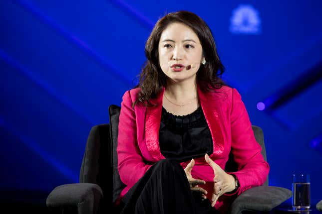 a woman wearing a bright pink blazer over a black shirt speaks while wearing a mic and sitting in a grey chair in front of a blue backdrop