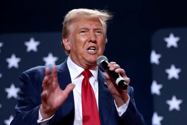 Donald Trump speaking in to a mic in his left hand, his right hand is up, he is standing in front of a navy background with white stars