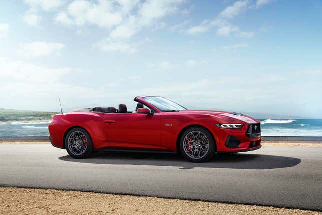 Red, convertible 2024 Ford Mustang on black pavement near a sandy beach.