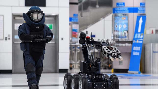 A police officer wearing full clear is seen with a remote control. A bomb disposal robot moves in front of them.