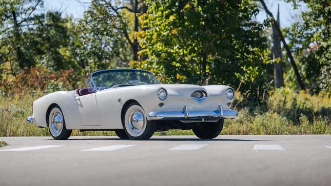 Un Kaiser Darrin blanc avec le toit baissé, garé dans une rue devant des arbres