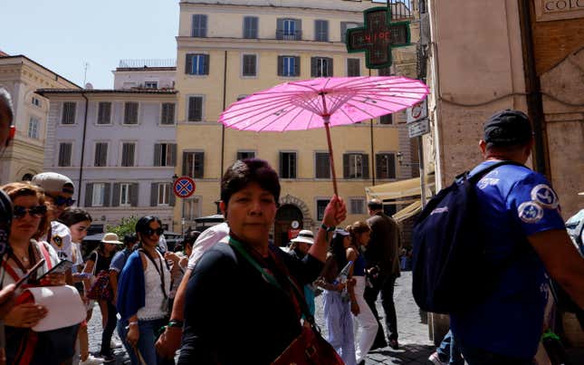 A woman with brown skin and short black hair holds a pink umbrella to shade from the sun, while standing in a Roman square in front of a yellow building. 