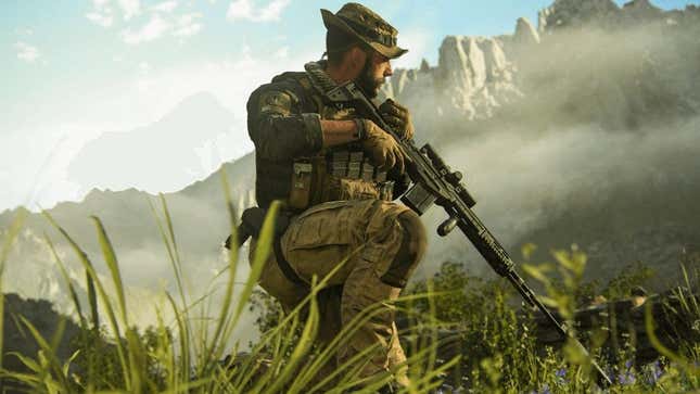 A soldier crouches in a grassy area with mountains in the background.