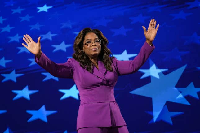 Opray with both her arms up wearing a purple suit in front of a blue backdrop with stars