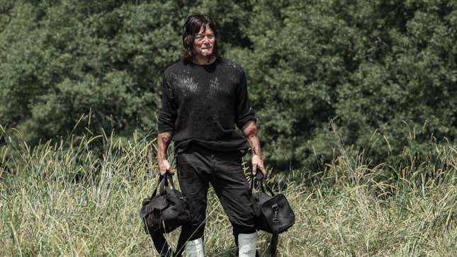 Daryl, wearing a filthy black sweatshirt, holds two black duffel bags while in a field.