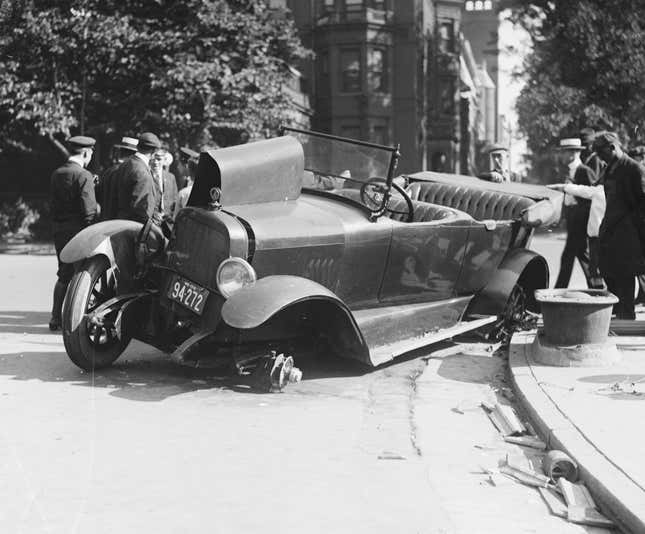 A crowd views the scene of an automobile accident, early twentieth century. The car shows extensive damage and rests on its left side, with both wheels on that side gone