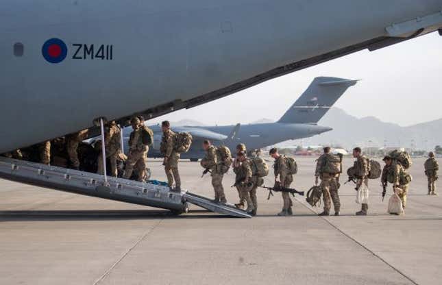 UK military personnel board an A400M aircraft departing Kabul. Some carry rifles, all carry bags, and all wear fatigues as they line up single file to walk up the ramp and into the plane, with mountains in the distance.