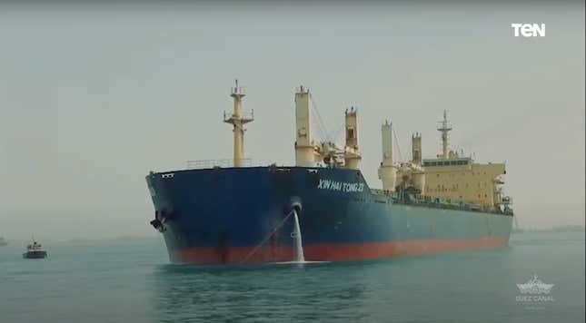 A 635-foot ship sailing through the Suez Canal. It's a hazy day, the ship is dark blue on the top and red at the waterliine with beige deck structures. A small tug boat appears alongside of it. 