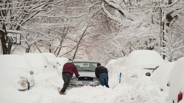 A photo of two people pushing a car out a snowdrift. 