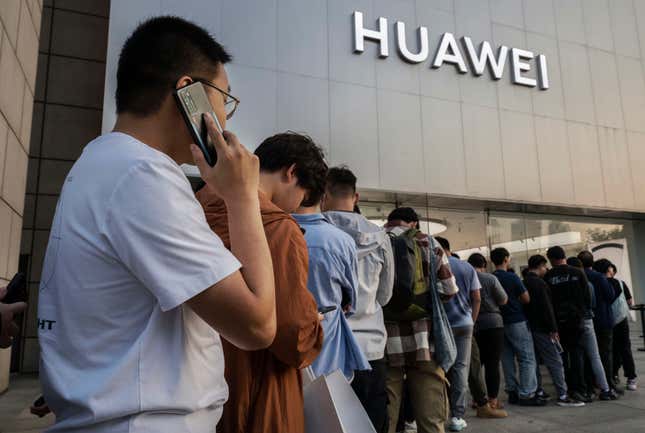 A man wearing a white t shirt talks on a Huawei mobile phone while waiting in line in front of a Huawei store