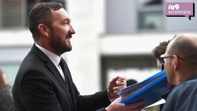 Ralph Ineson signing an autographs while wearing a black suit and smiling at the crowd.