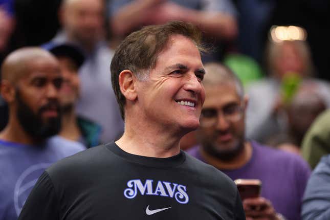 Mark Cuban porte un maillot noir avec le logo des Mavs et un motif Nike souriant et regardant vers la gauche