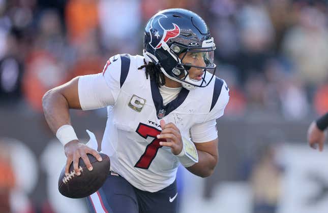 A man with long locks, a navy blue helmet, and a white Houston Texans jersey with red number palms the football while running on an NFL football field. 