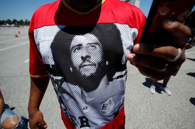 A Black man wears a t-shirt with Colin Kaepernick's face on it while holding a phone.
