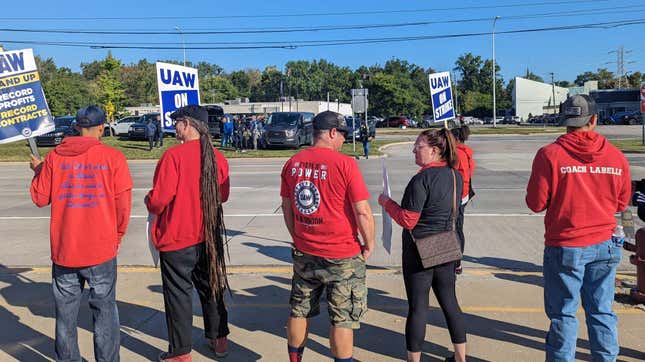 Six people all wearing red and holding UAW ON STRIKE signs stand along a four lane road on a sunny day