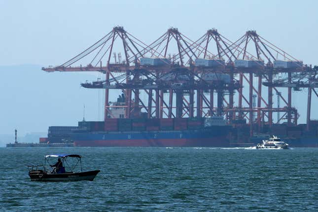 A man on a boat fishes as a ferry sails by a containers vessel docked at a port shrouded by haze in Xiamen in southeast China&#39;s Fujian province on Dec. 26, 2023. The United Nations issued a somber global economic forecast for 2024 on Thursday, Jan. 4, 2024, pointing to challenges from escalating conflicts, sluggish global trade, persistently high interest rates and increasing climate disasters. (AP Photo/Andy Wong)