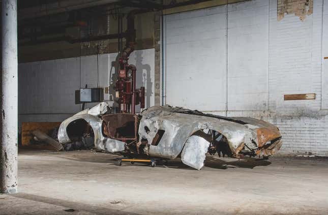 The burned out and rusty wreckage of a 1954 Ferrari 500 Mondial Spider Series I in a depressing looking basement. The white painted brick walls are chipping and there's some kind of brass utilities pipes behind it. 