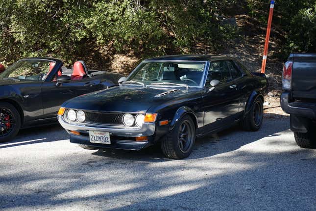 A dark blue 1972 Toyota Celica is parked in the shadows next to a late model Miata.