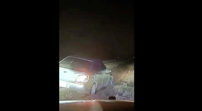 A car in the grassy median of a highway illuminated by a police cruiser's headlights. 