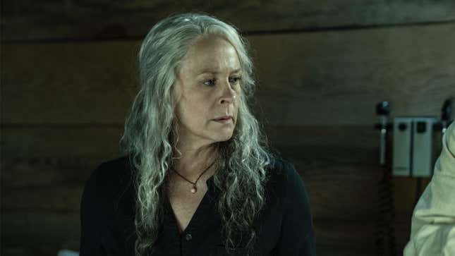 Carol, with long silver hair half-pulled into a tail, talks to someone in a wood-paneled room.