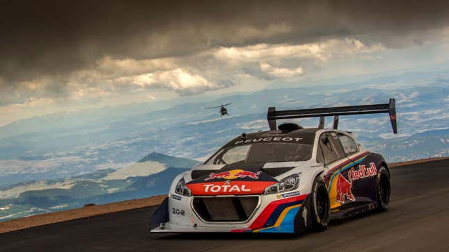 A photo of a Peugeot car racing up the Pikes Peak Hillclimb course with a helicopter in the background. 