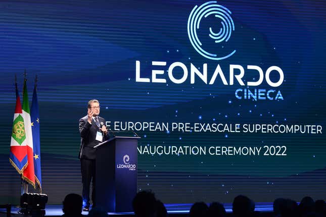 a man standing at a podium on stage in front of three flags and a backdrop that says LEONARDO CINECA