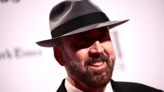Nic Cage, in a black suit and gray fedora that shades his eyes, smiles at the crowd.