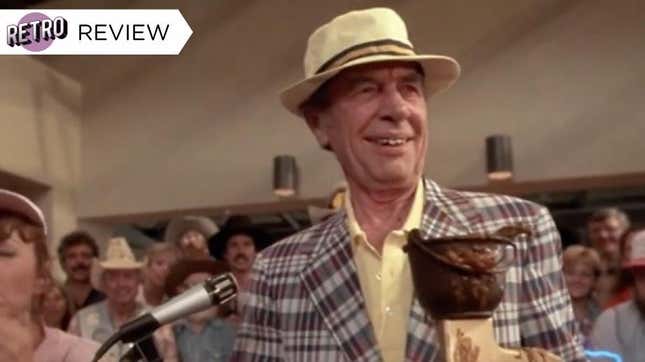 A man in a tan, brimmed hat and plaid jacket holds his first-prize chili cook-off trophy as he stands before a crowd and a microphone.